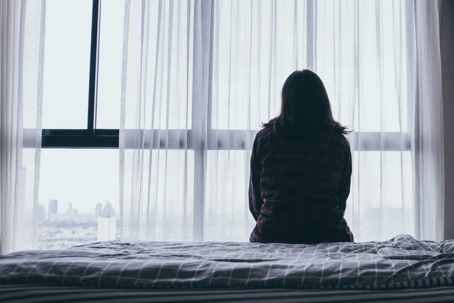 Woman sitting alone on bed looking out window.