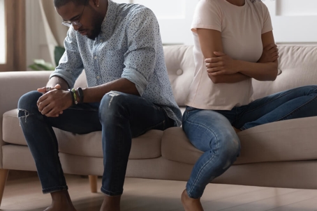 Couple sits on couch facing in opposite directions. The man on the left has his hands in his lap while looking down and the woman has her arms crossed looking away.