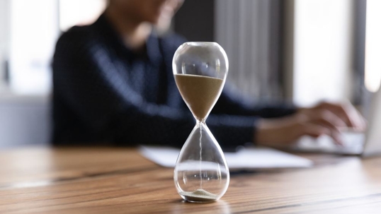 Close up view of hourglass stand on wooden home office measuring time, woman busy using computer.