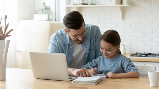 Father sitting next to daughter at table with computer and notebook.
